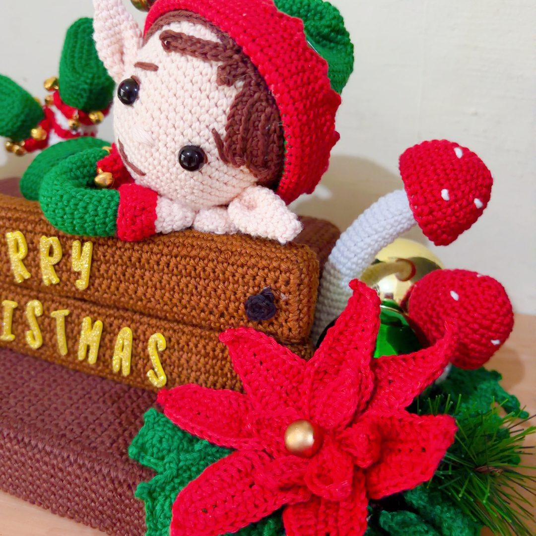 crochet elf on crochet wood, surrounded by poinsettias, holly leaves, mushrooms and a lantern
