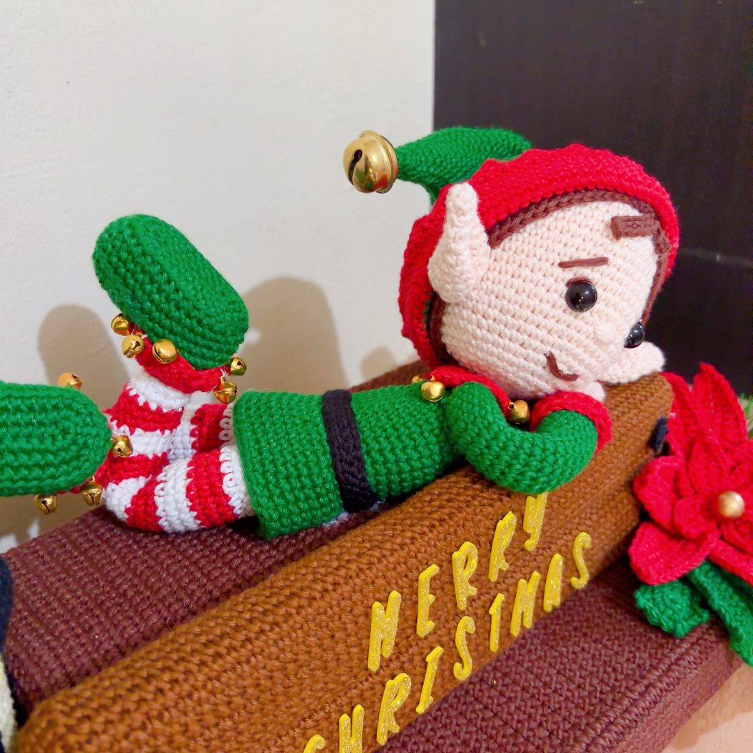 crochet elf on crochet wood, surrounded by poinsettias, holly leaves, mushrooms and a lantern