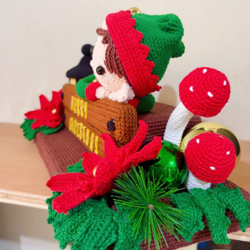 duende de crochê sobre uma peça de madeira feita em crochê, cercado por poinsétias, folhas de azevinho, cogumelos e uma lanterna