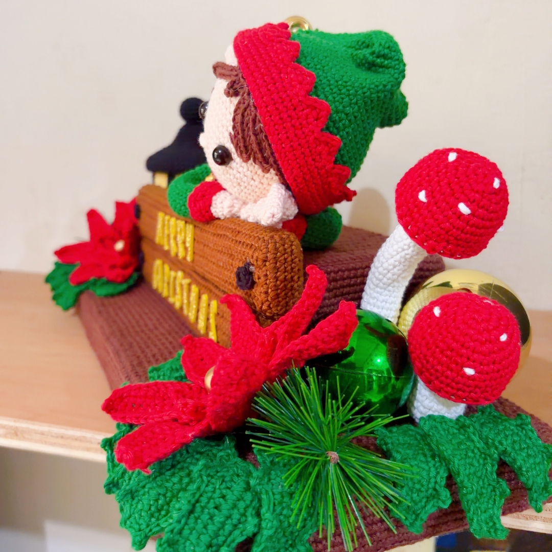 crochet elf on crochet wood, surrounded by poinsettias, holly leaves, mushrooms and a lantern