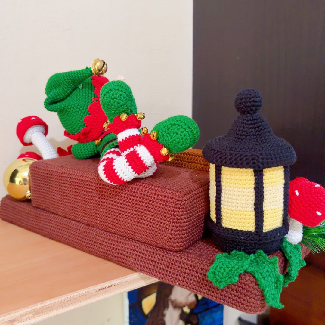 crochet elf on crochet wood, surrounded by poinsettias, holly leaves, mushrooms and a lantern