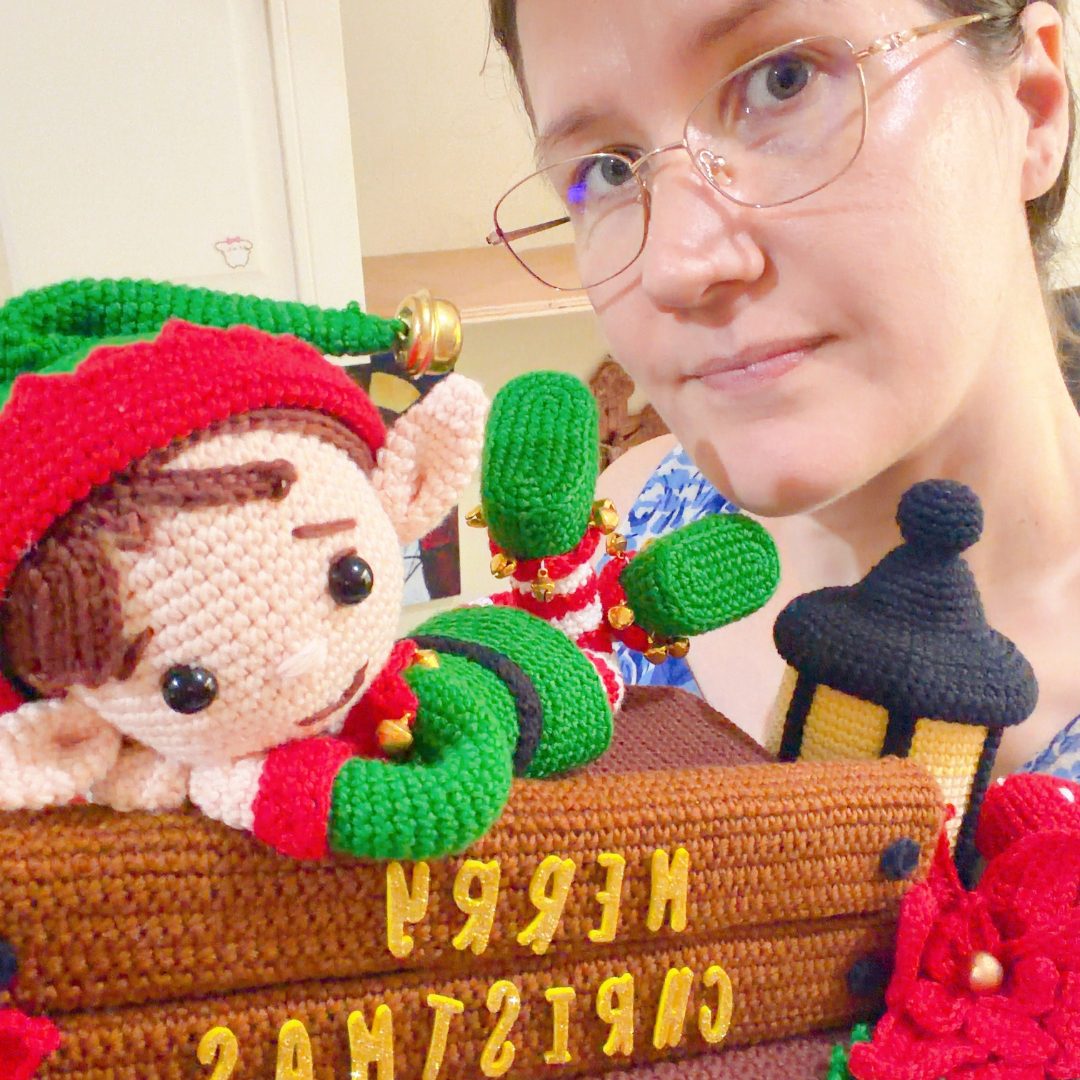 crochet elf on crochet wood, surrounded by poinsettias, holly leaves, mushrooms and a lantern
